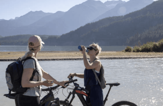 Two women with electric bikes by a lake, enjoying a break, mountains in the background